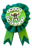 A ribbon made of green and light green felt, with the character Shrek glued onto it and beads that spell out 'swamp dweller' in all-caps.